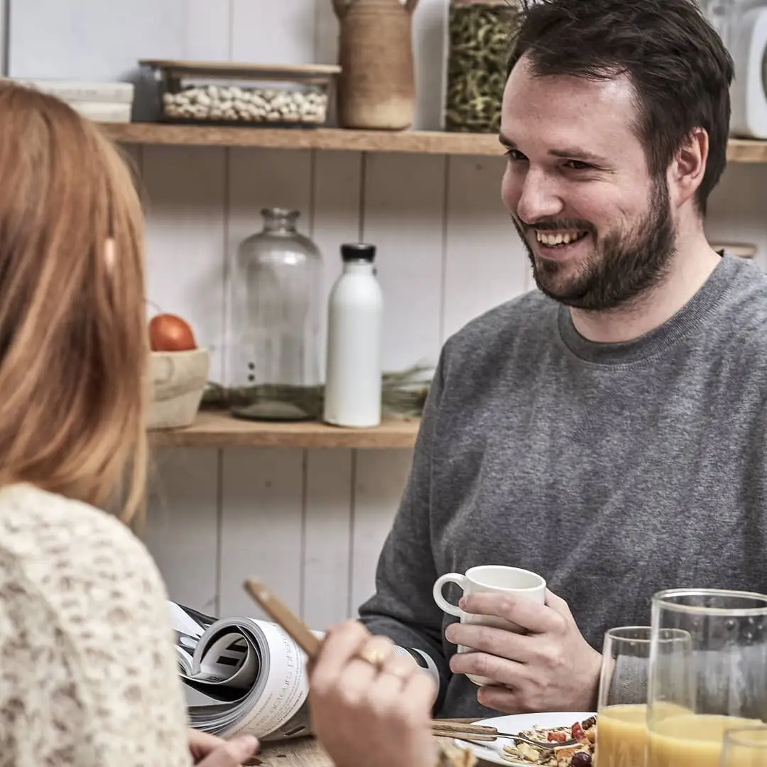Lohan, souriant, tenant une tasse, partageant un moment convivial autour d'un petit-déjeuner avec une femme dont on ne voit que le dos.